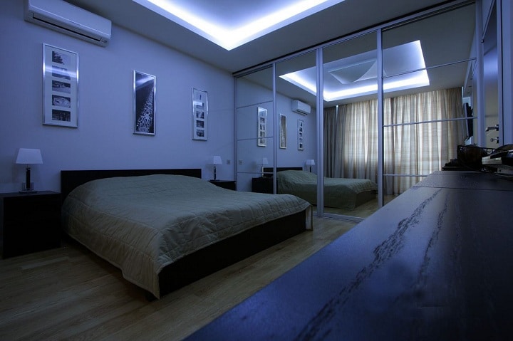 Спальня в светлом цвете, в качестве декора используются картины, пано на стене над кроватью. Центральное освещение дублируется светодиодной лентой, плюс настольные светильники. Шкаф в интерьере встроен в нишу. Потолок плотно примыкает к дверям, для расширения пространства используются зеркала.
