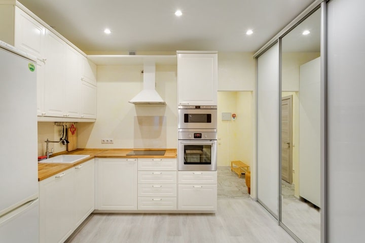 Белая матовая кухня с рифленым гарнитуром под классическую отделку. Верхние шкафы сделаны только по одной стене. При входе размещена стойка с необходимым оборудованием для приготовления пищи.